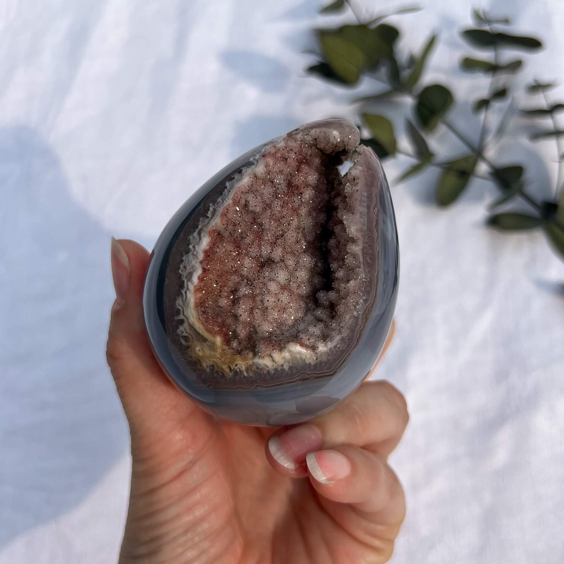 A hand holding a polished amethyst crystal geode egg with a soft blue outer shell and a druzy pink core against a white background