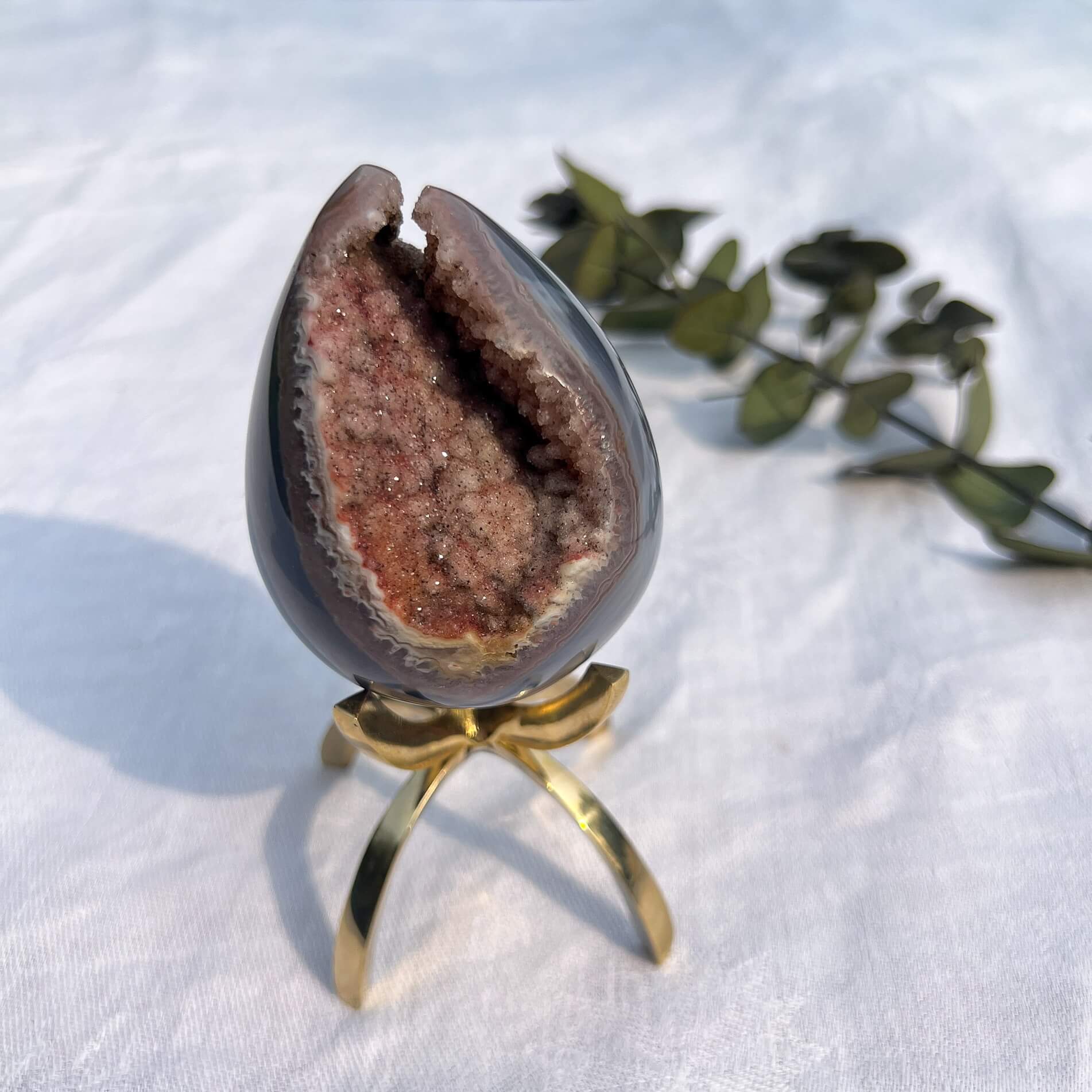 A polished amethyst crystal geode egg with a soft blue outer shell and a druzy pink core, displayed on a gold-colored stand.