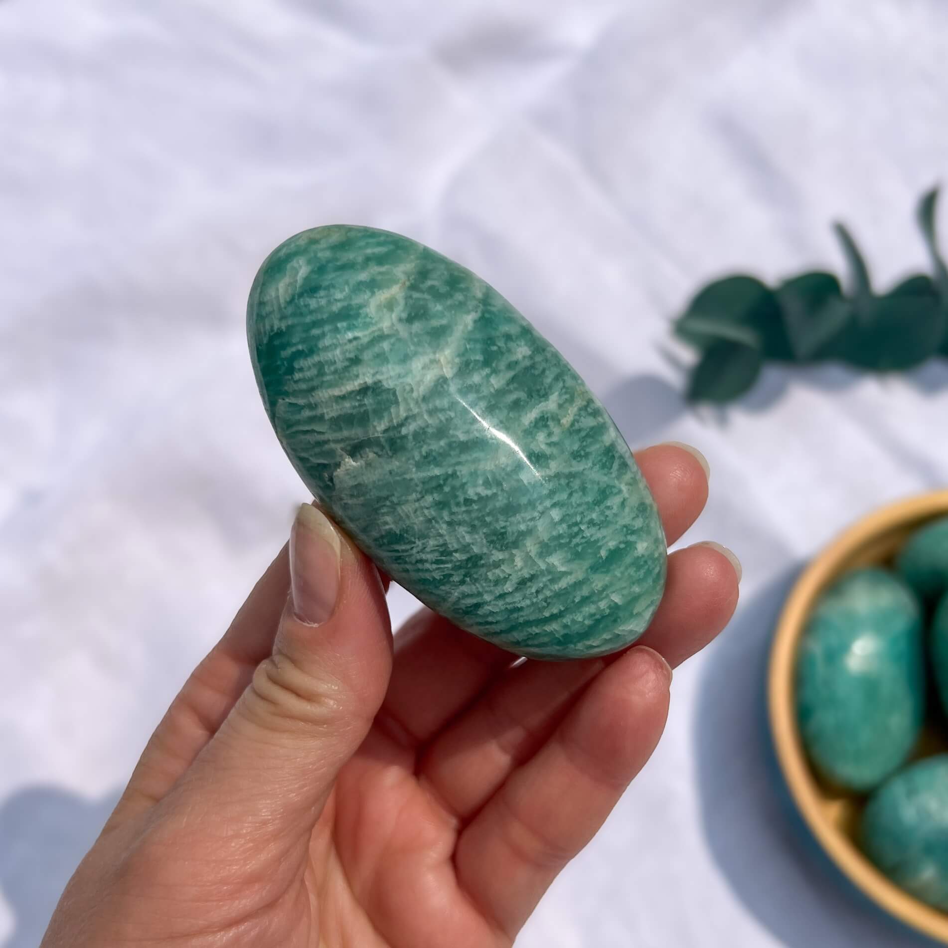Hand holding a turquoise and white striped amazonite crystal palmstone with a wooden dish of green palmstones in the background