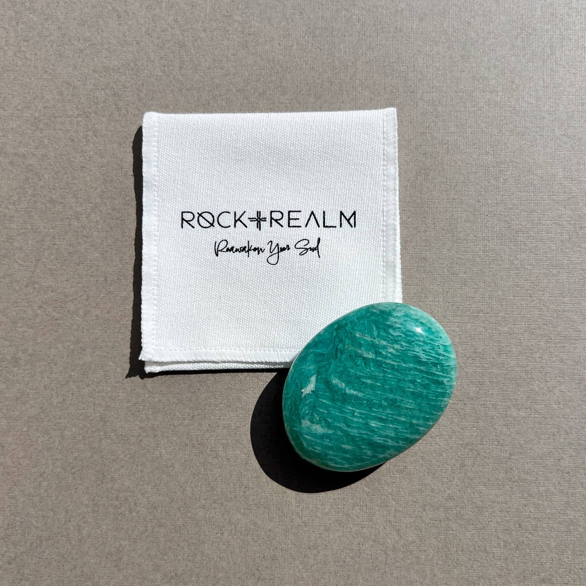 A polished oval-shaped turquoise-green amazonite crystal palm stone next to a white cotton pouch printed with the brand name 'ROCK + REALM' set against a neutral taupe background.