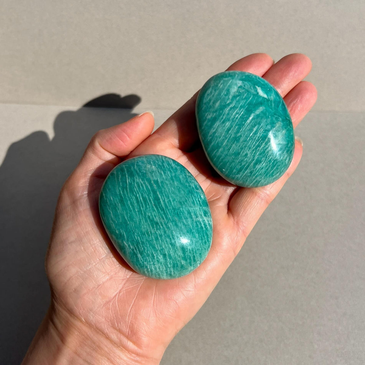Two polished turquoise amazonite crystal palmstones with a marbled pattern, held in a person's hand against a neutral grey background.