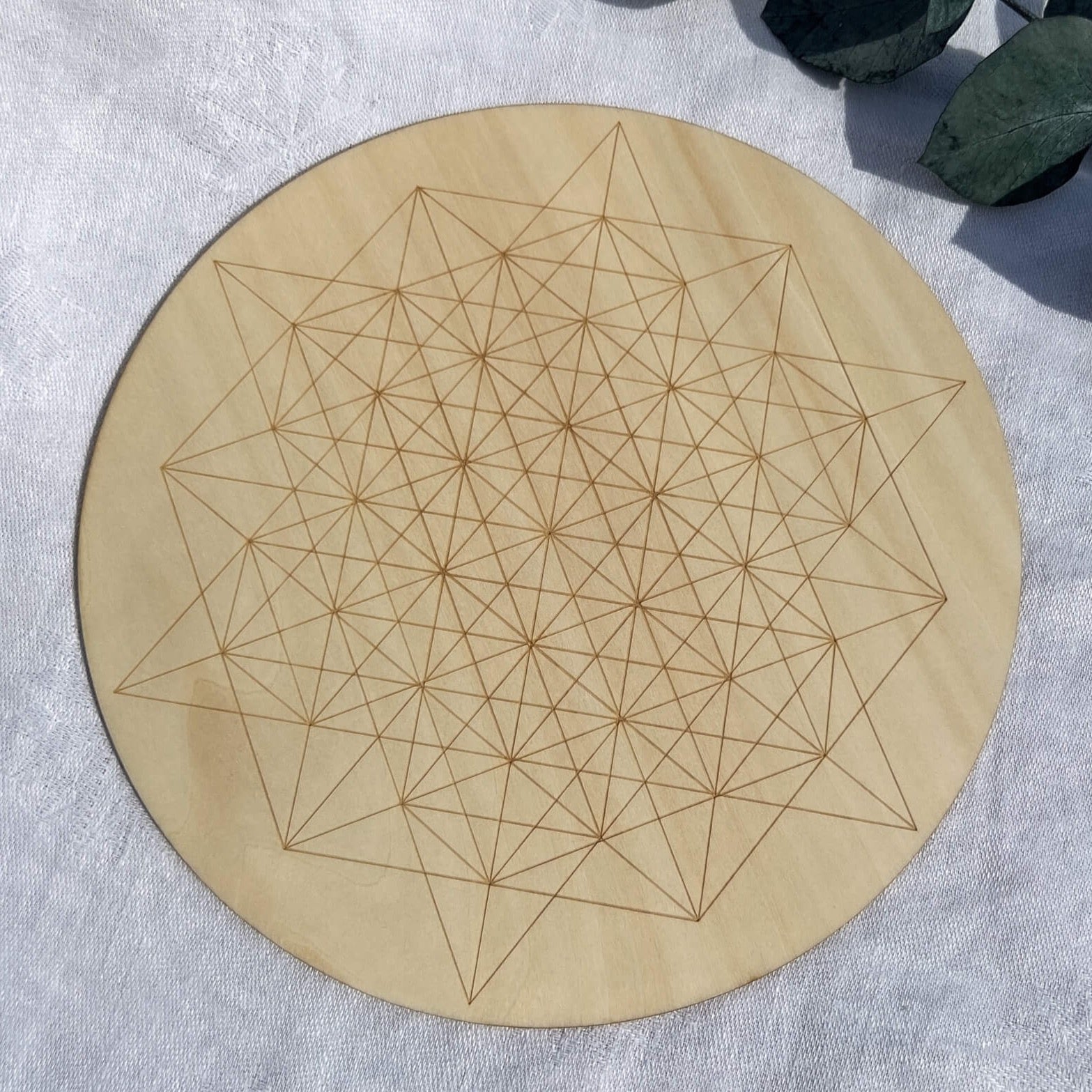 A wooden crystal grid board with an etched geometric ascension grid design, placed on a white surface with a green leaf in the corner.