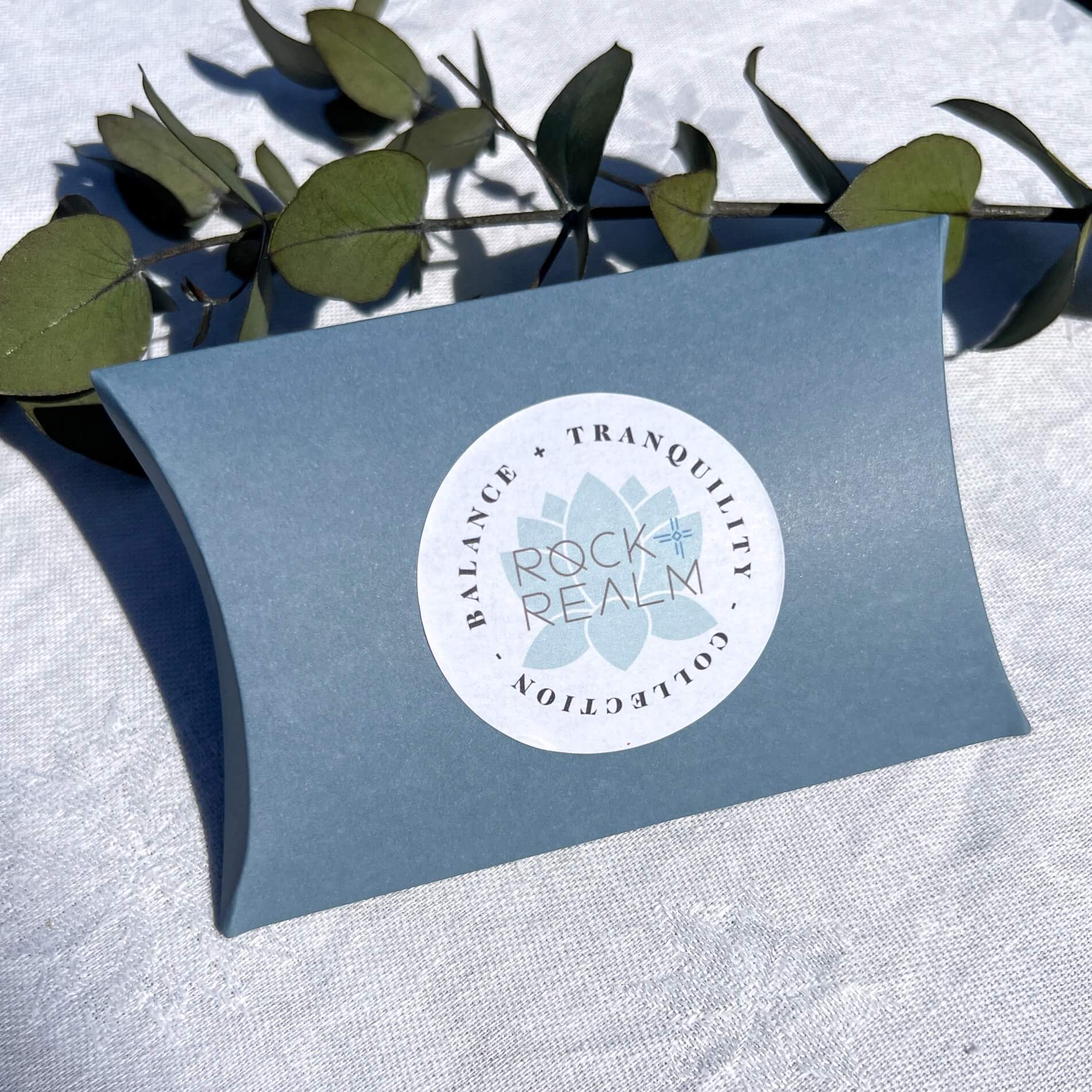 A pale blue pillow box with a white and green label stating 'Balance + Tranquility Collection by Rock + Realm' against a white background with green leaves