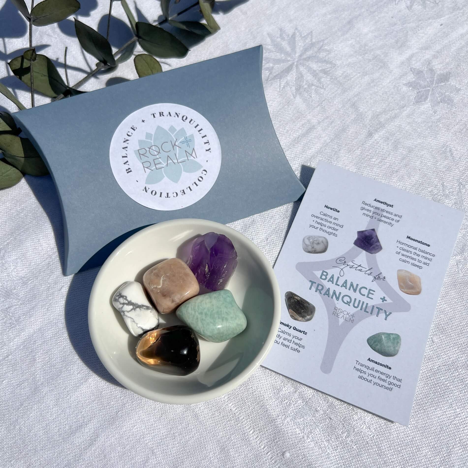A collection of various crystals placed in a white dish with a crystal properties card next to it, all presented on a white fabric surface.
