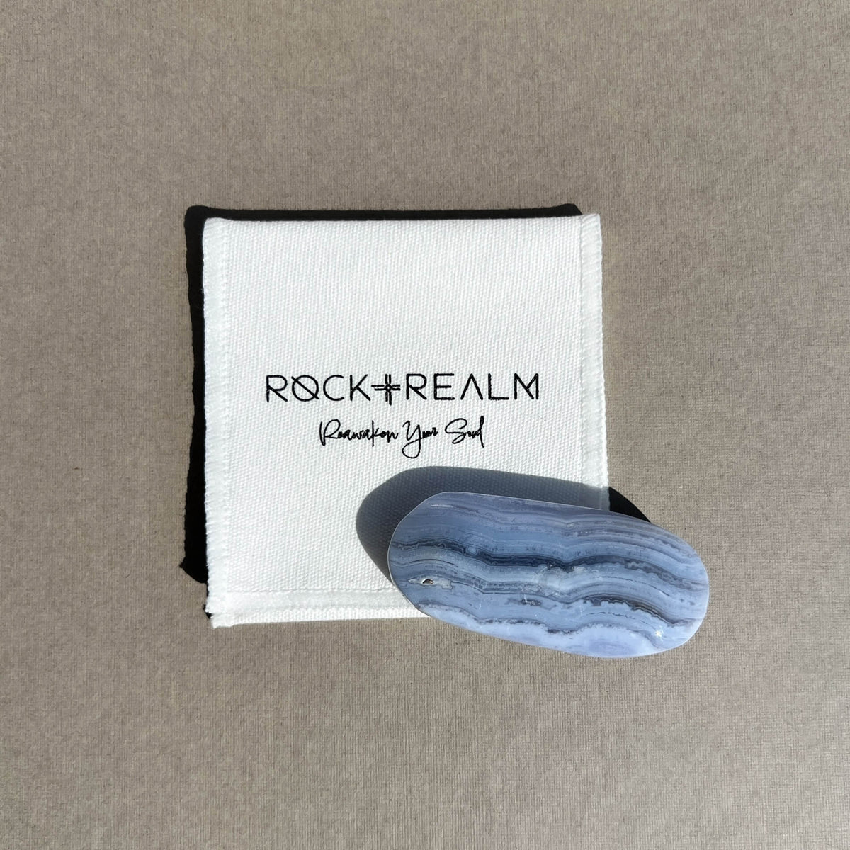 A polished oval blue lace agate palmstone with natural white banding, displayed on top of a white cotton branded 'ROCK + REALM Reawaken Your Soul' against a neutral taupe background.