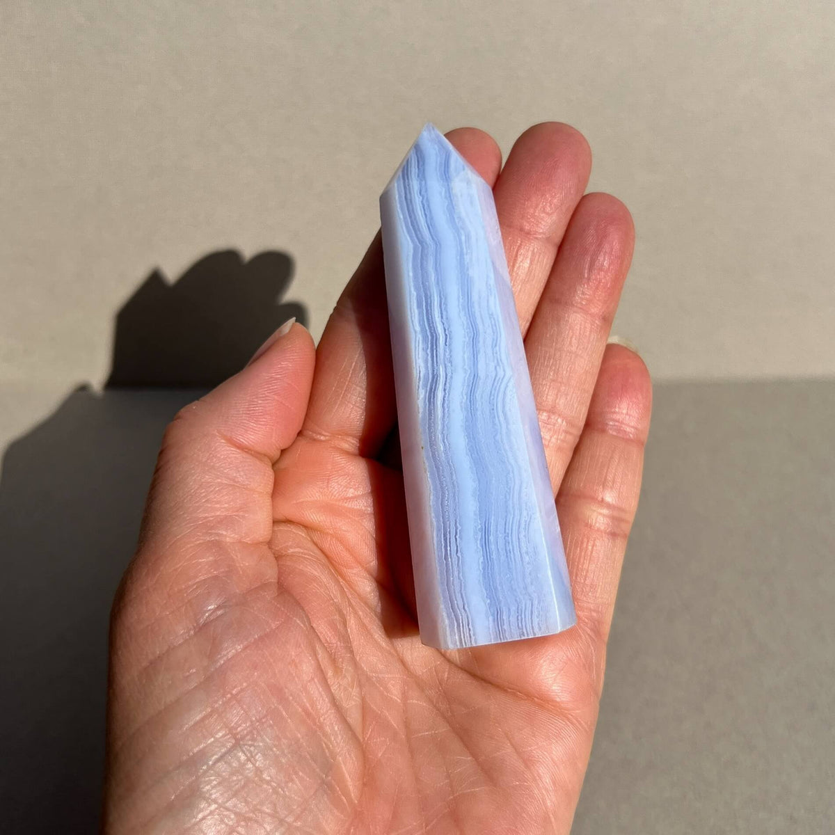 A hand holding a polished light blue lace agate crystal point against a neutral beige background with a soft shadow.
