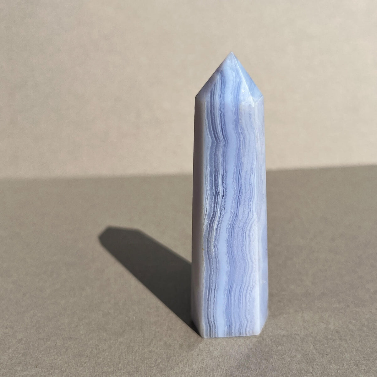 A polished blue and white banded blue lace agate crystal tower with a pointed tip, standing upright on a neutral gray surface against a beige background.