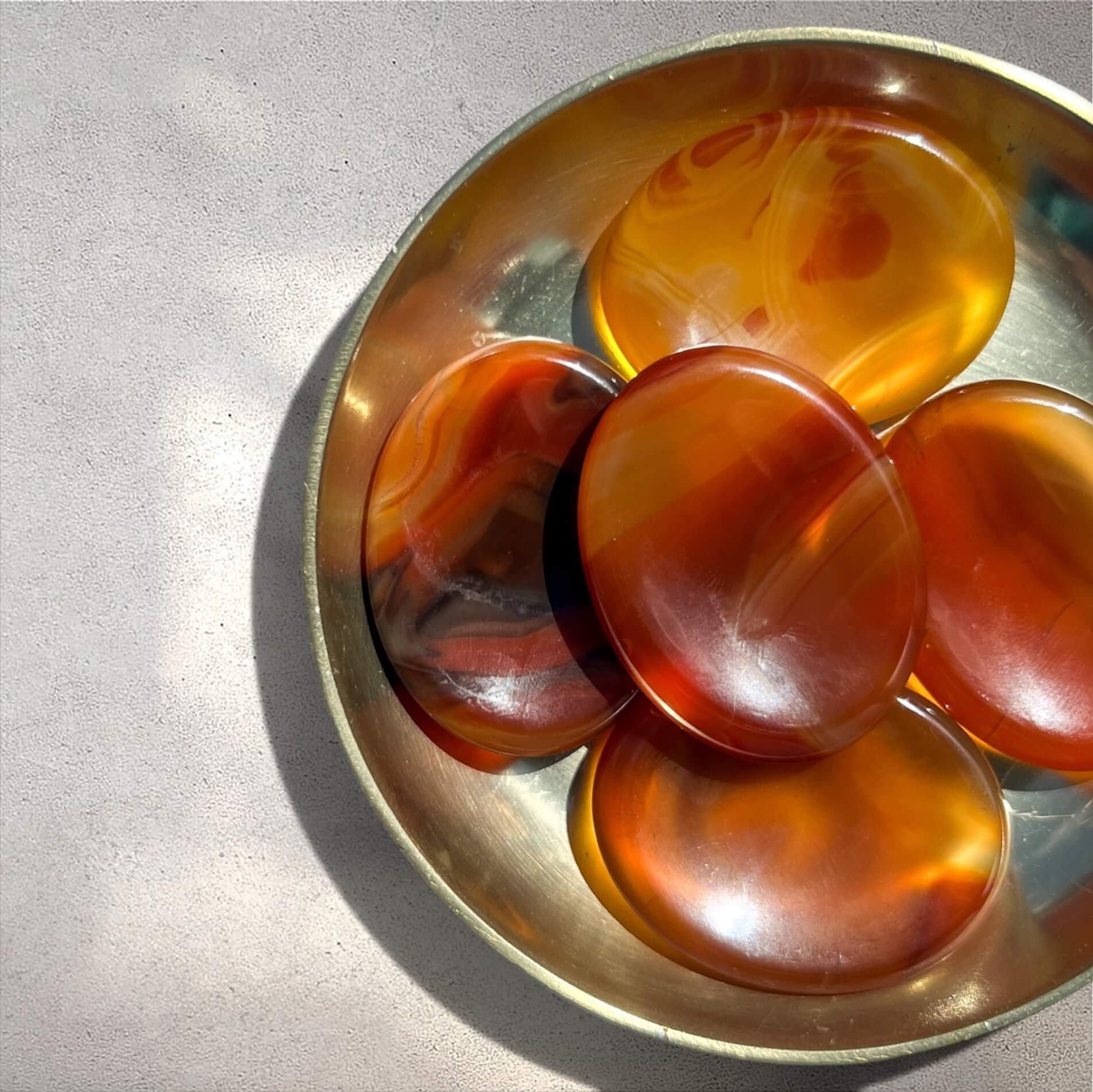 Five polished oval carnelian crystal worrystones with amber, orange and red marbled patterns arranged in a small brass dish on a light grey surface.