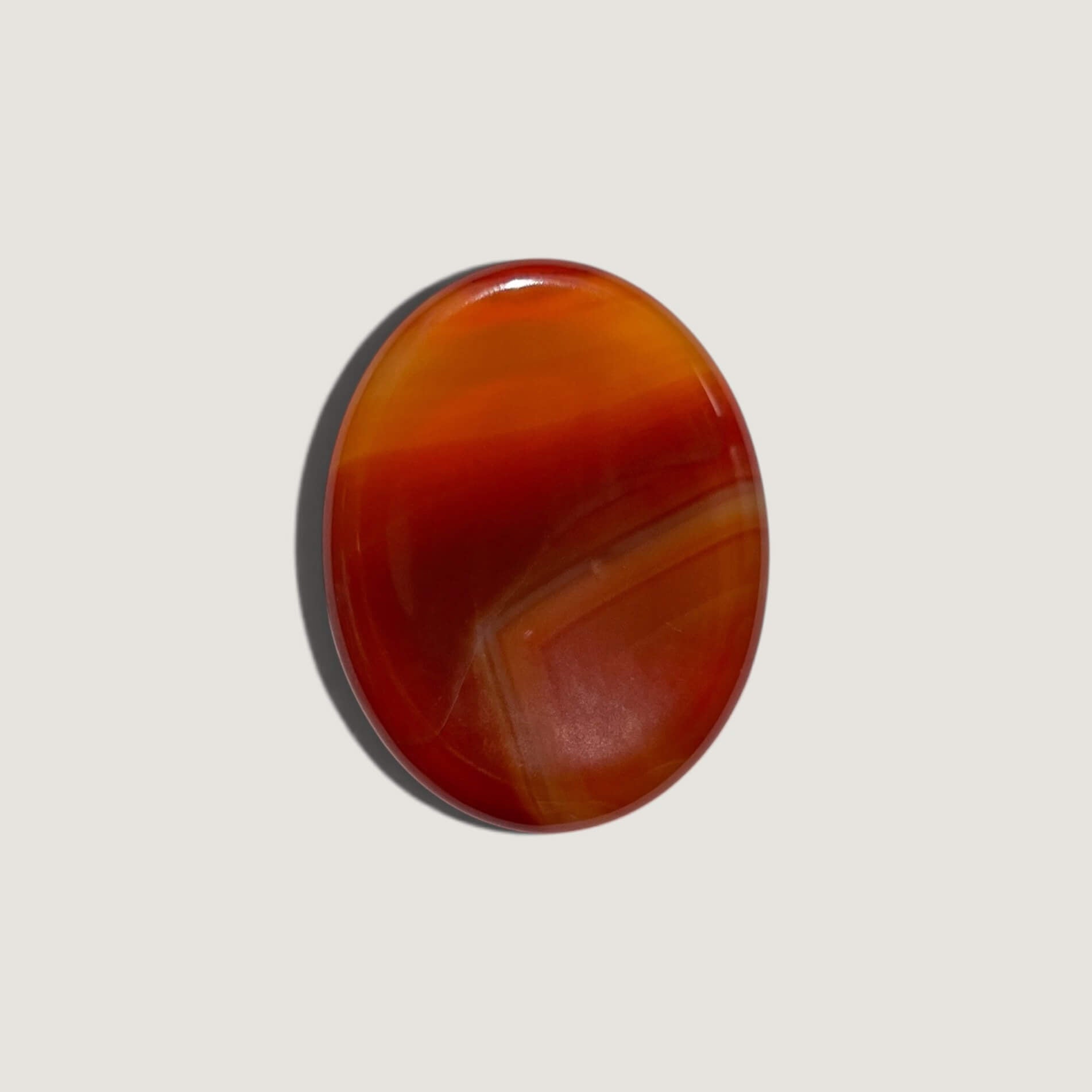 A polished oval carnelian worry stone with natural banding in shades of orange, red and brown, displayed on a light grey background with a soft drop shadow.