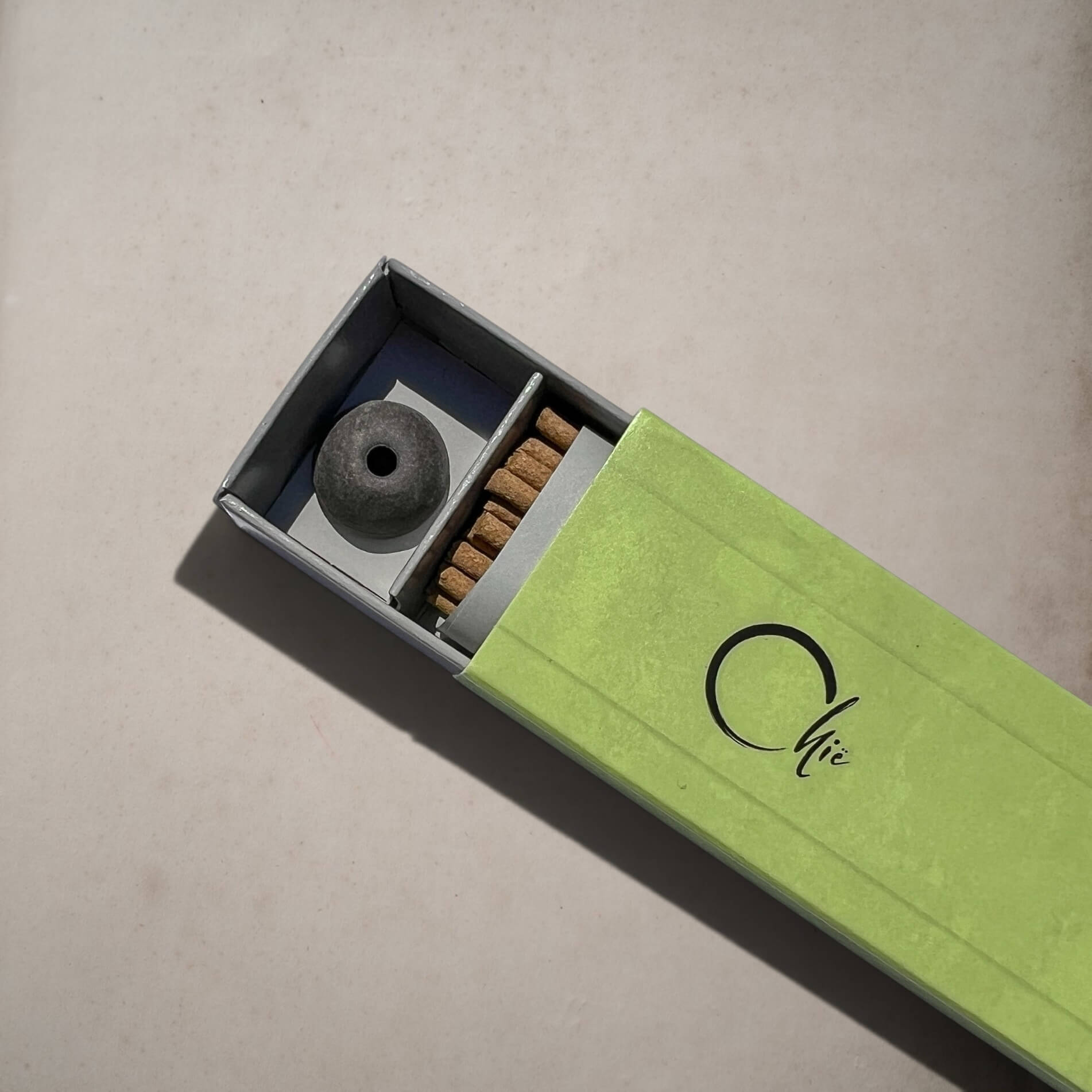 A light green matchbox-style incense box with a sliding lid, shown open to reveal a round dark incense holder and several incense sticks. The lid has a black script logo reading 'Chie'.