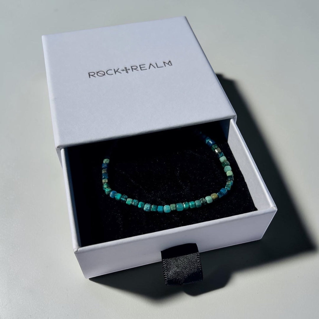 Blue, green, aqua and teal coloured Chrysocolla crystal cube bead bracelet in a 'Rock + Realm' box on a white fabric background