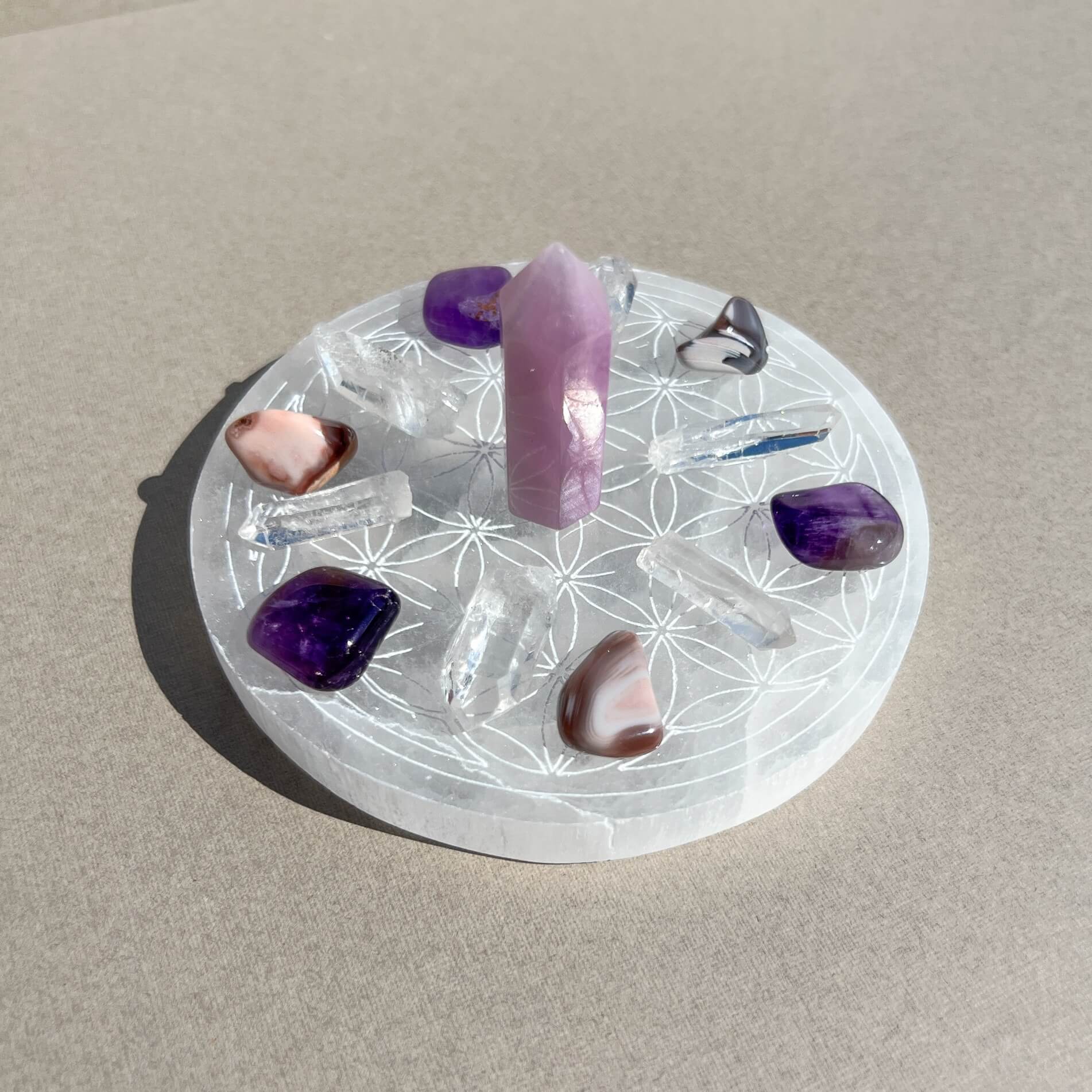 A circular white selenite crystal plate with etched geometric patterns, holding several polished crystals including a kunzite point, dark purple amethyst and agate tumbles and clear quartz points, photographed on a neutral beige surface.