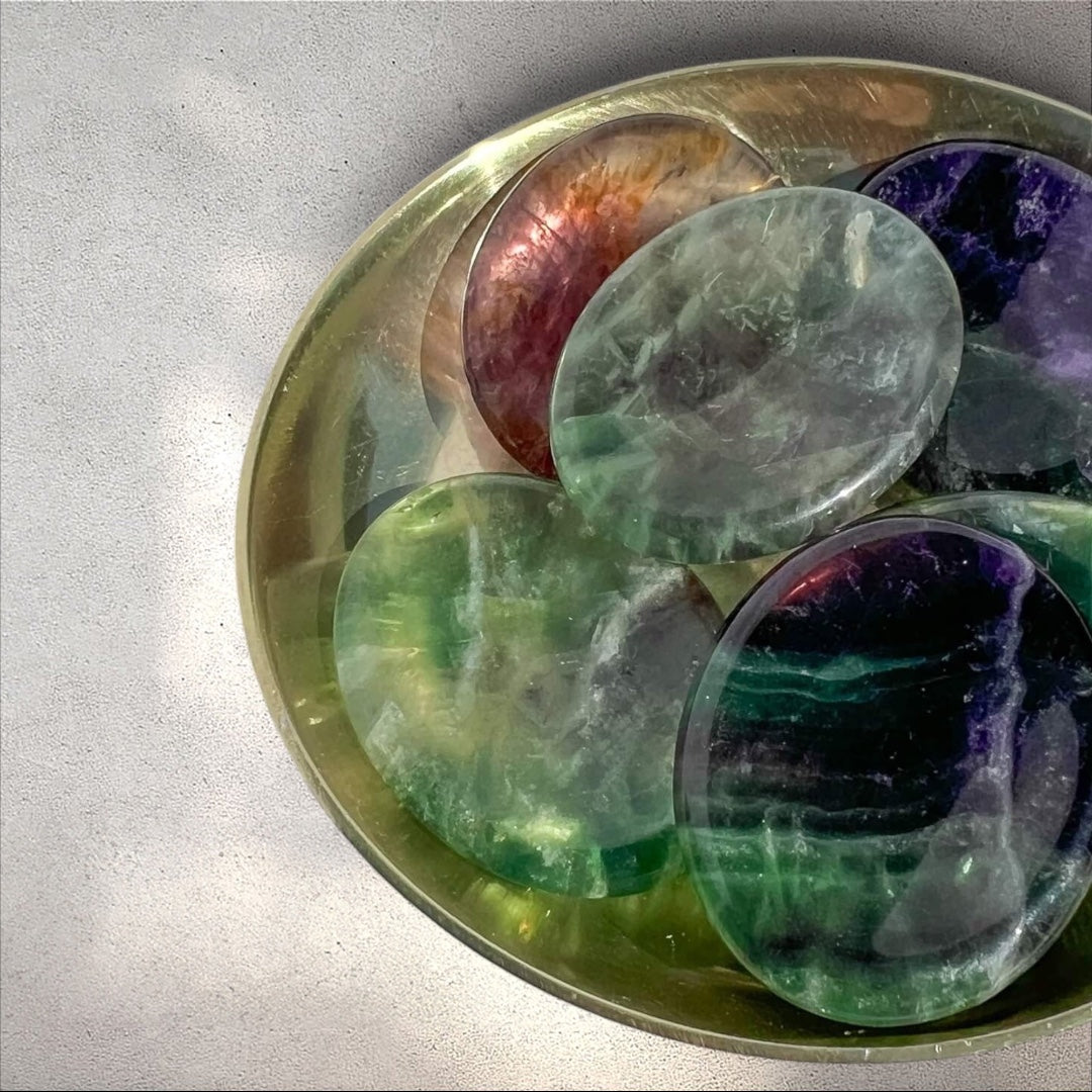 Brass dish full of polished fluorite worry stones in green, purple, and pink hues, oval-shaped with a thumb indentation.