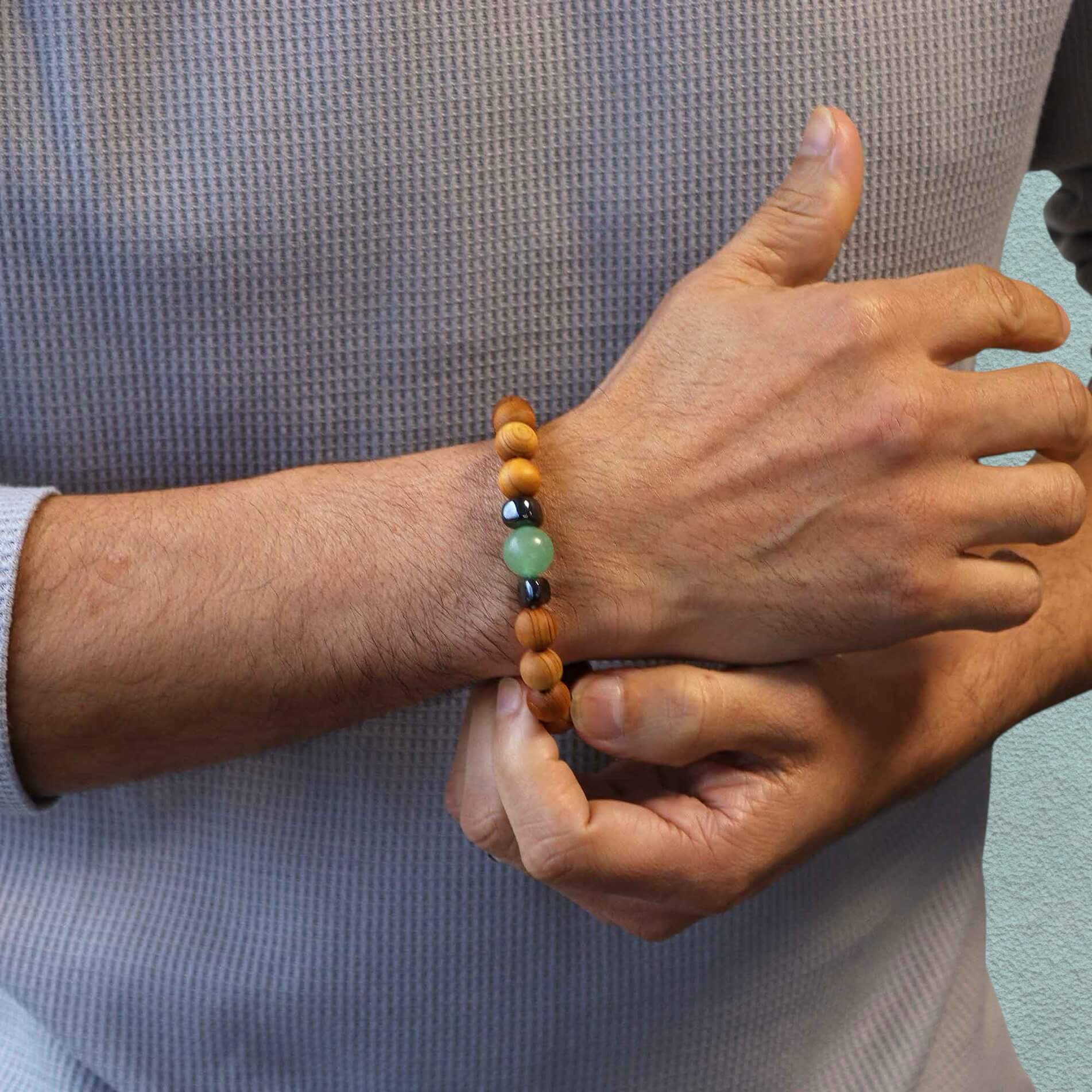 Man in grey top wearing a Cedar wood beaded bracelet with green aventurine and metallic black hematite beads stood against a neutral background