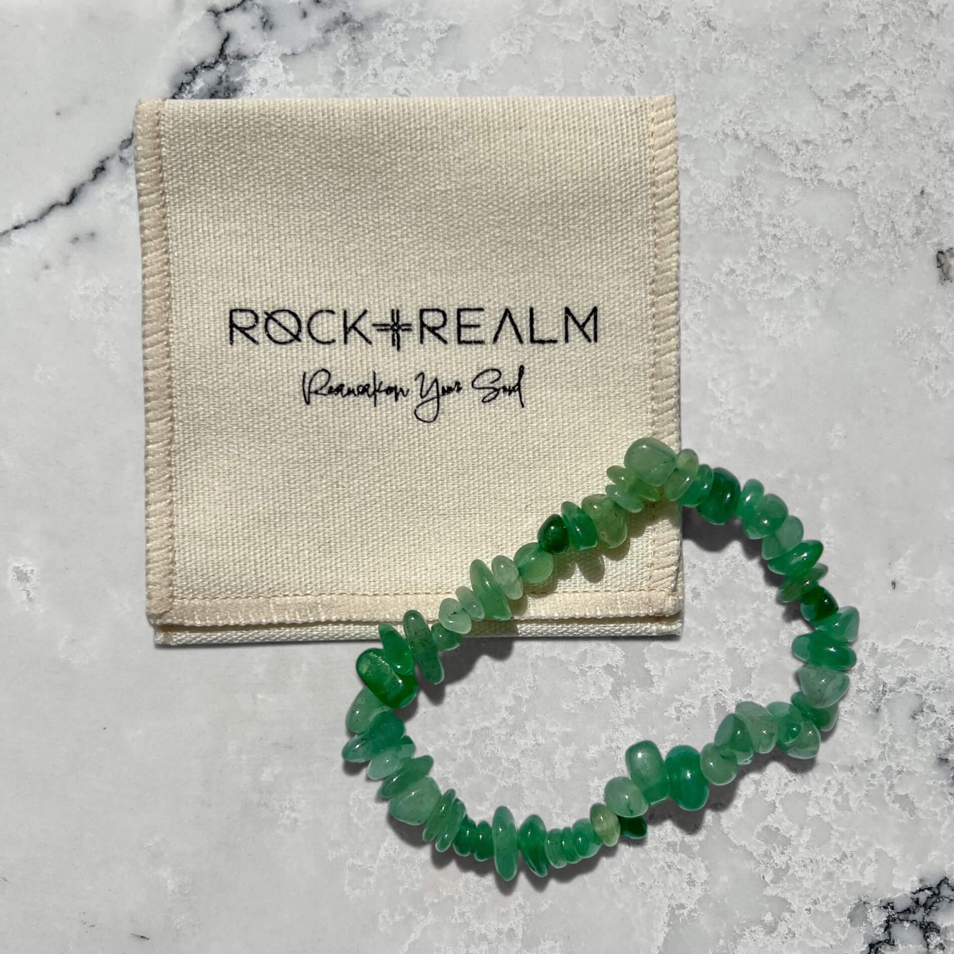 Green Aventurine crystal chip bracelet on a marble surface with 'Rock + Realm' cotton pouch.
