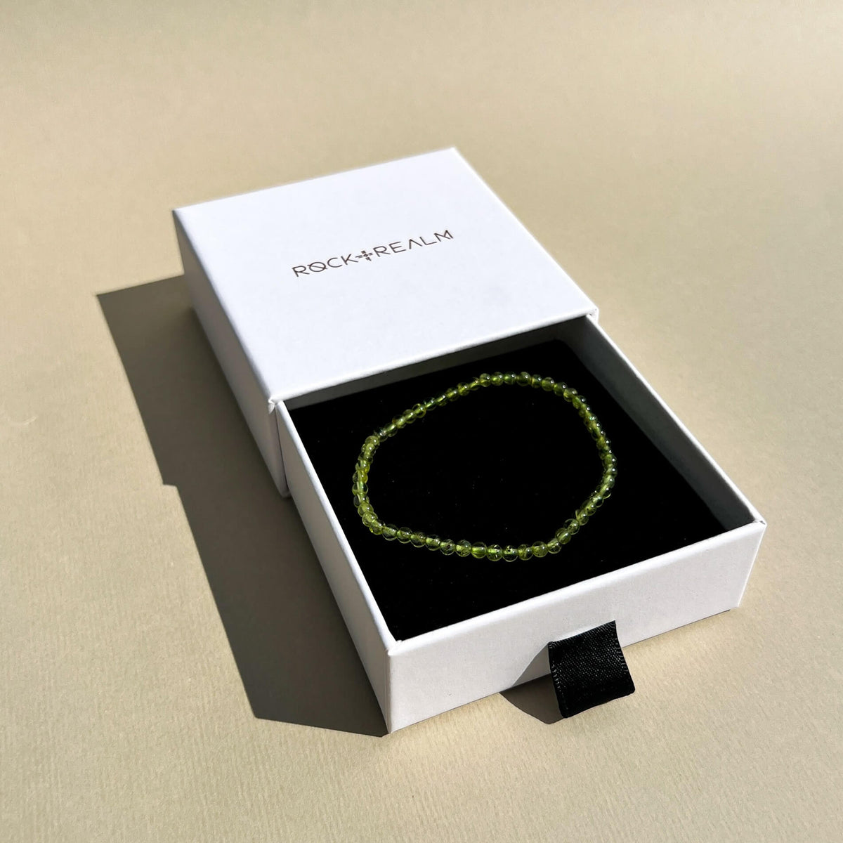 A green Peridot crystal beaded bracelet displayed inside an open white ROCK + REALM jewellery box with a black insert, set on a beige textured surface.
