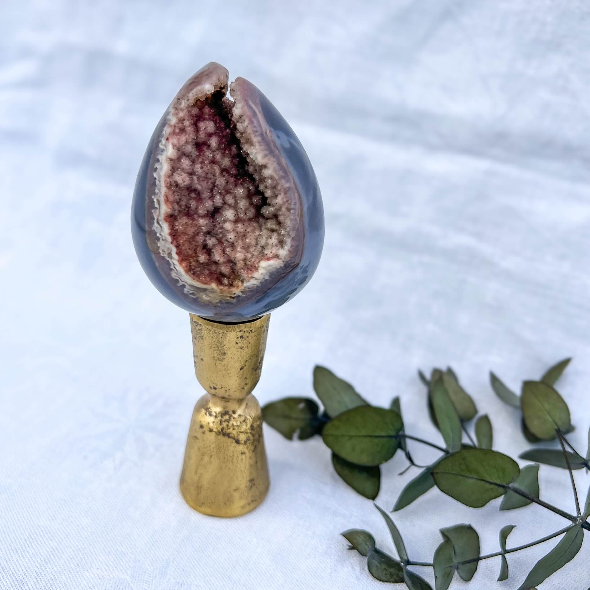 A polished amethyst crystal geode egg with a soft blue outer shell and a druzy pink core, displayed on a gold display stand against a white background with leaves