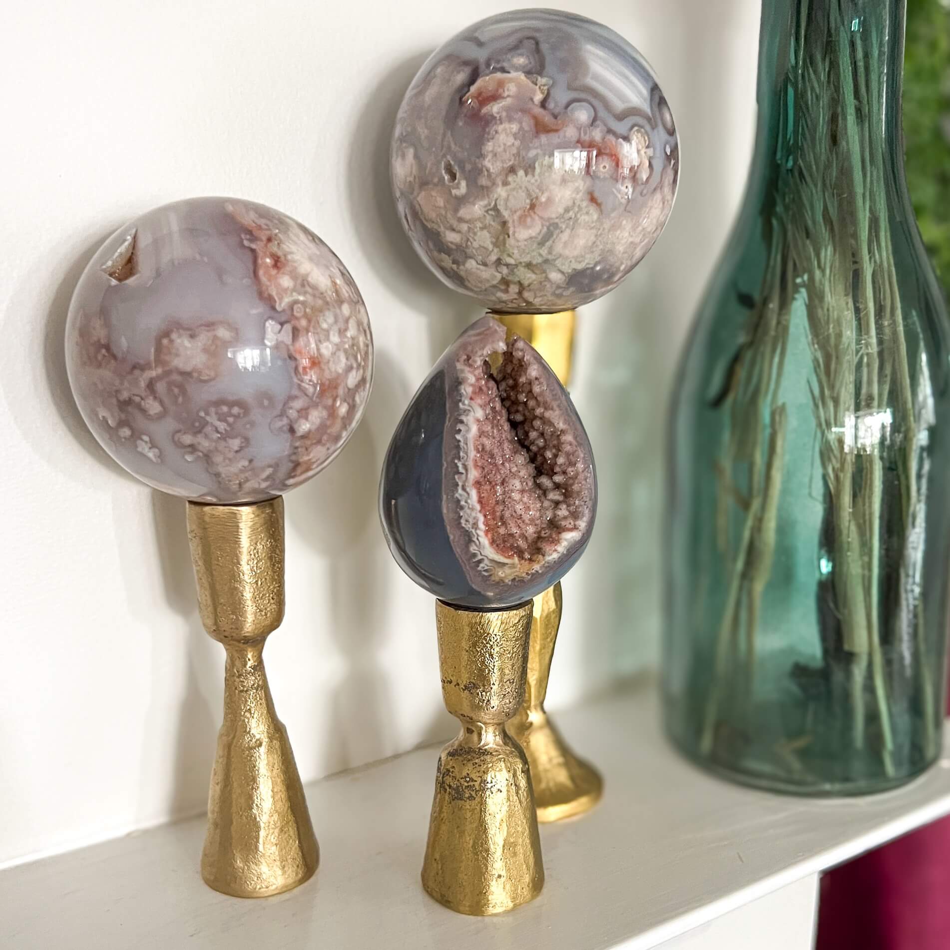 Three pink and blue polished display crystals on gold coloured stands on a mantlepiece next to a deep blue glass vase
