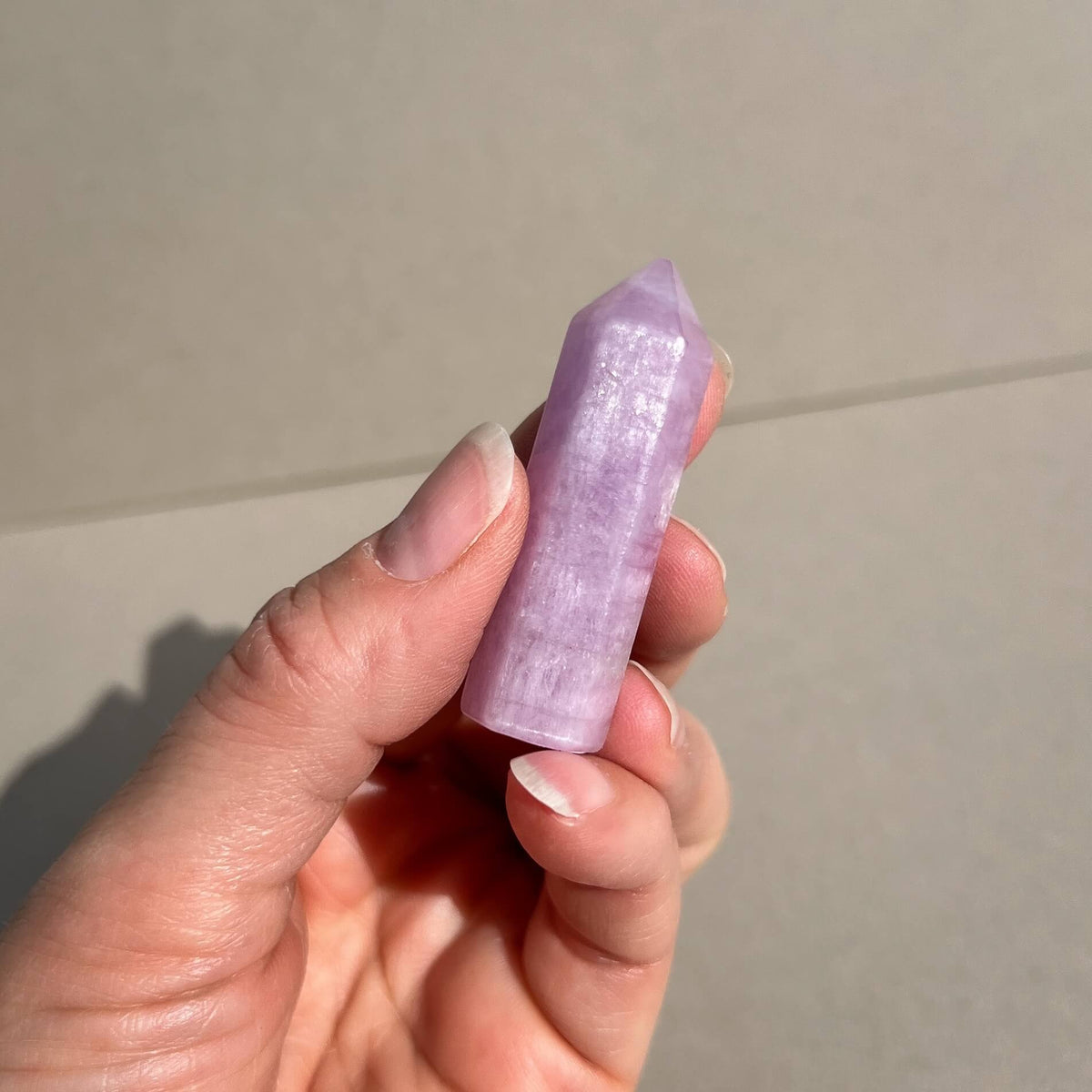 A hand holding a polished lavender-purple kunzite crystal point against a neutral beige background.