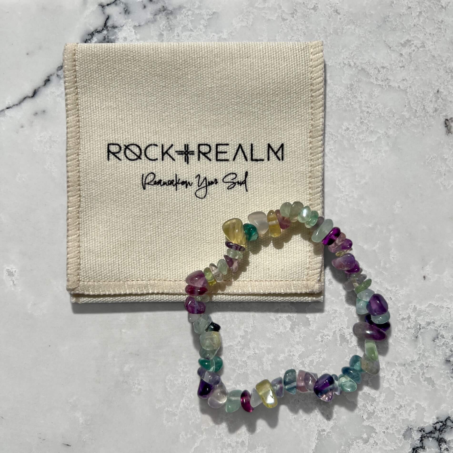 Multicolored rainbow fluorite gemstone chip bracelet on a marble surface with a cotton'Rock + Realm' pouch