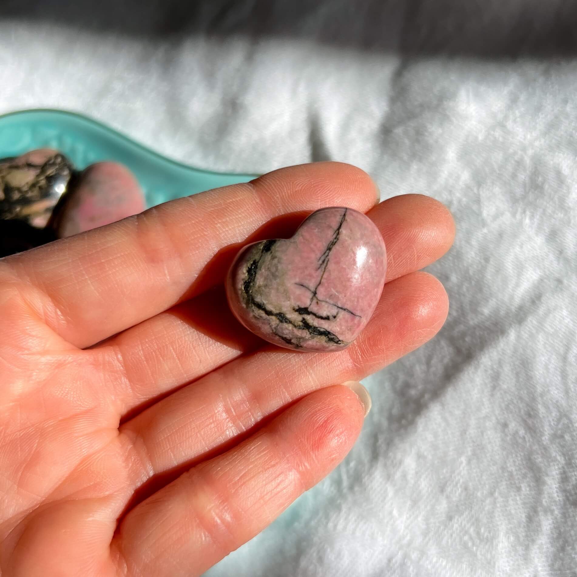 A hand holding a polished pink Rhodonite crystal heart with black patterns.