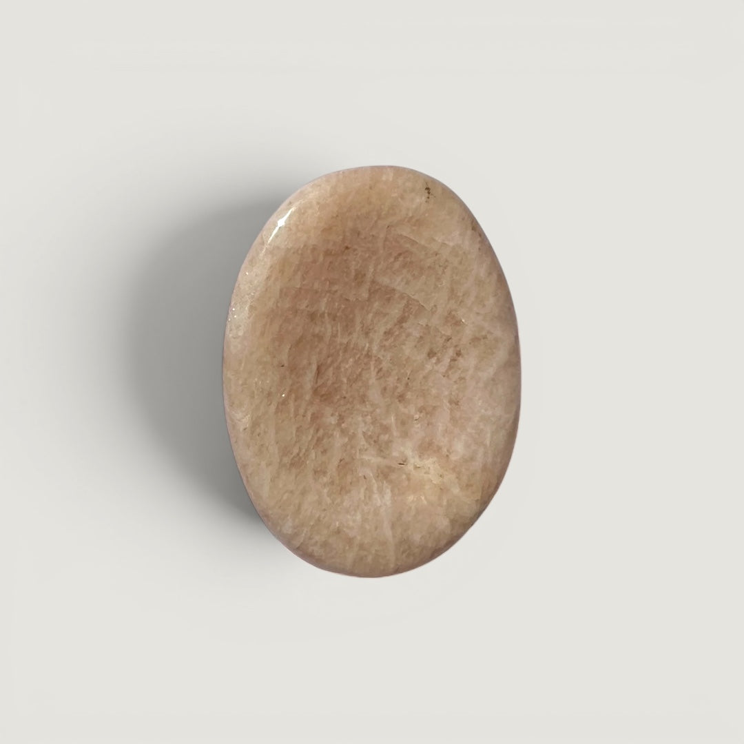 Polished oval peach moonstone worry stone with thumb indentation, beige with subtle shimmer.