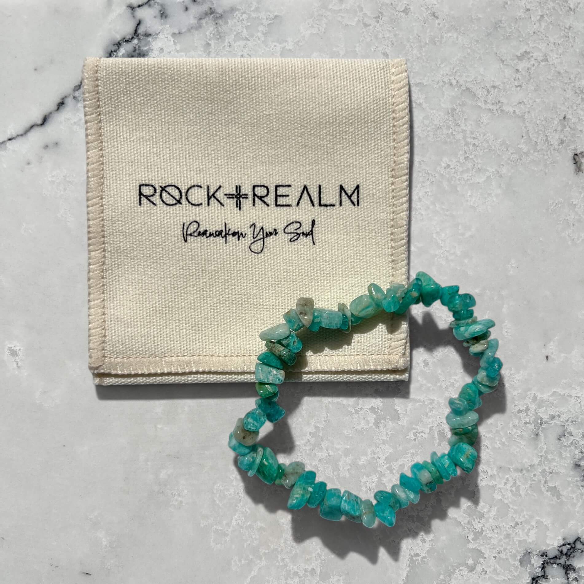 Turquoise Russian Amazonite crystal chip bracelet on a marble surface with 'Rock+Realm' branded cream cotton pouch