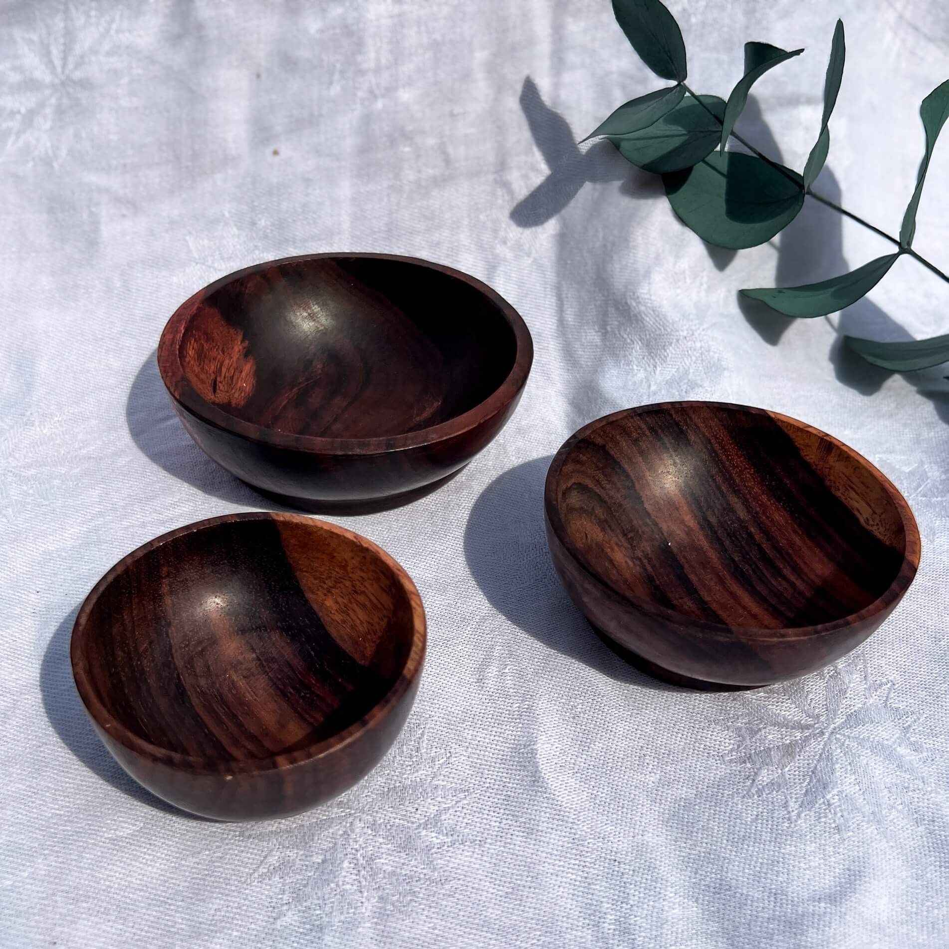 Three Indian Rose Wood bowls on a textured white surface with a leafy branch.