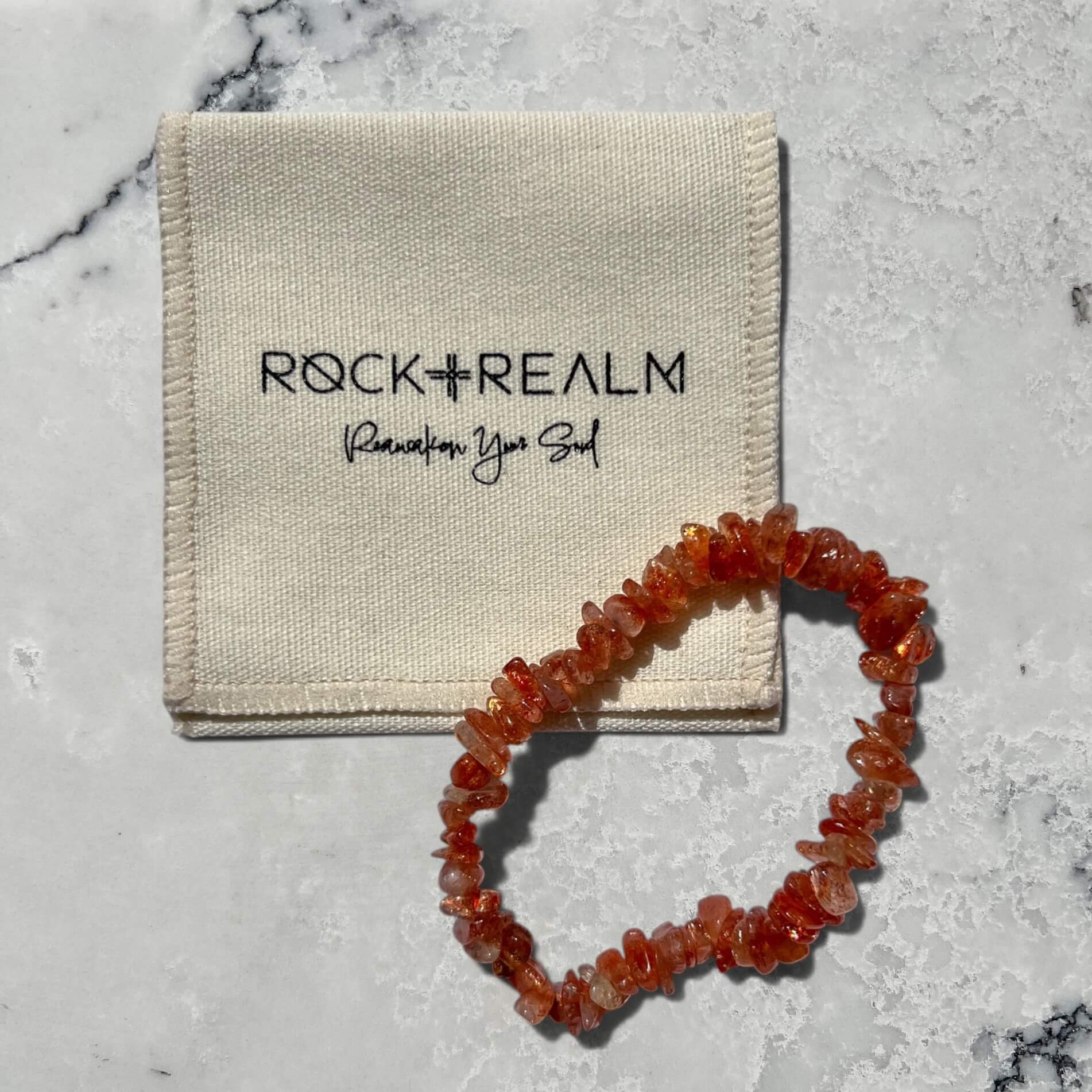 Orange high grade sunstone crystal chip bracelet with sparkle on a grey marble surface with cream cotton 'Rock + Realm' pouch.