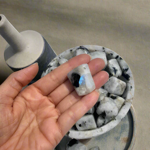 A woman's hand holding a large moonstone crystal with a blue flash with vase and bowl of crystals in the background