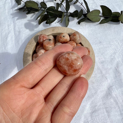 A large orange sunstone crystal tumblestone held to camera in an open palm with a dish of sunstone crystals in the background