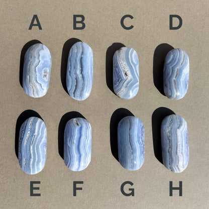 Eight polished oval blue lace agate palmstones labeled A through H are arranged in a 2x4 grid on a tan background. Each palmstone has a light blue, white and gray agate band pattern with darker veining.