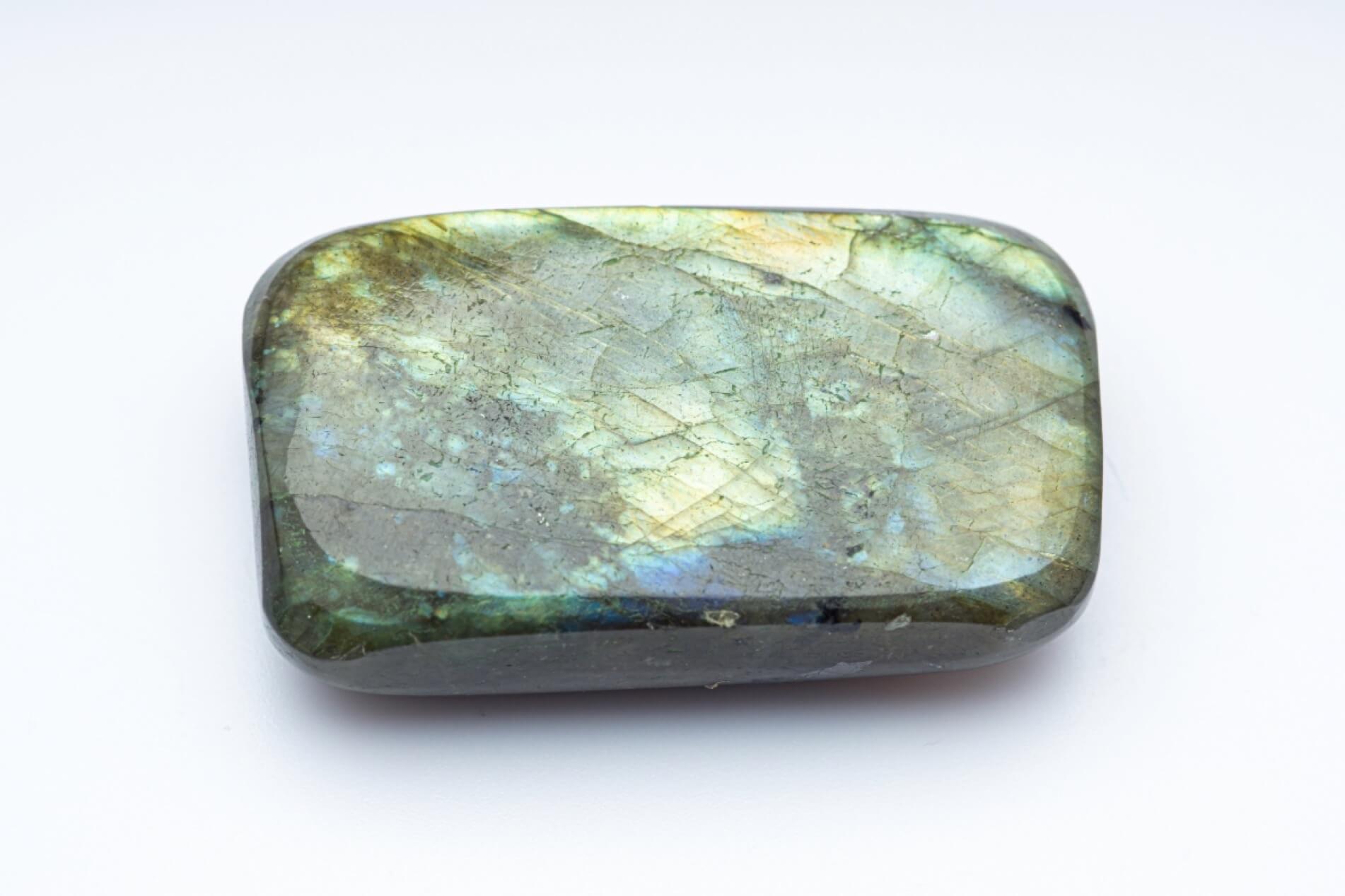 A polished labradorite stone with a rounded rectangular shape, displaying a shimmering iridescent play-of-color with green, blue, and gold hues over a gray base, set against a plain white background.