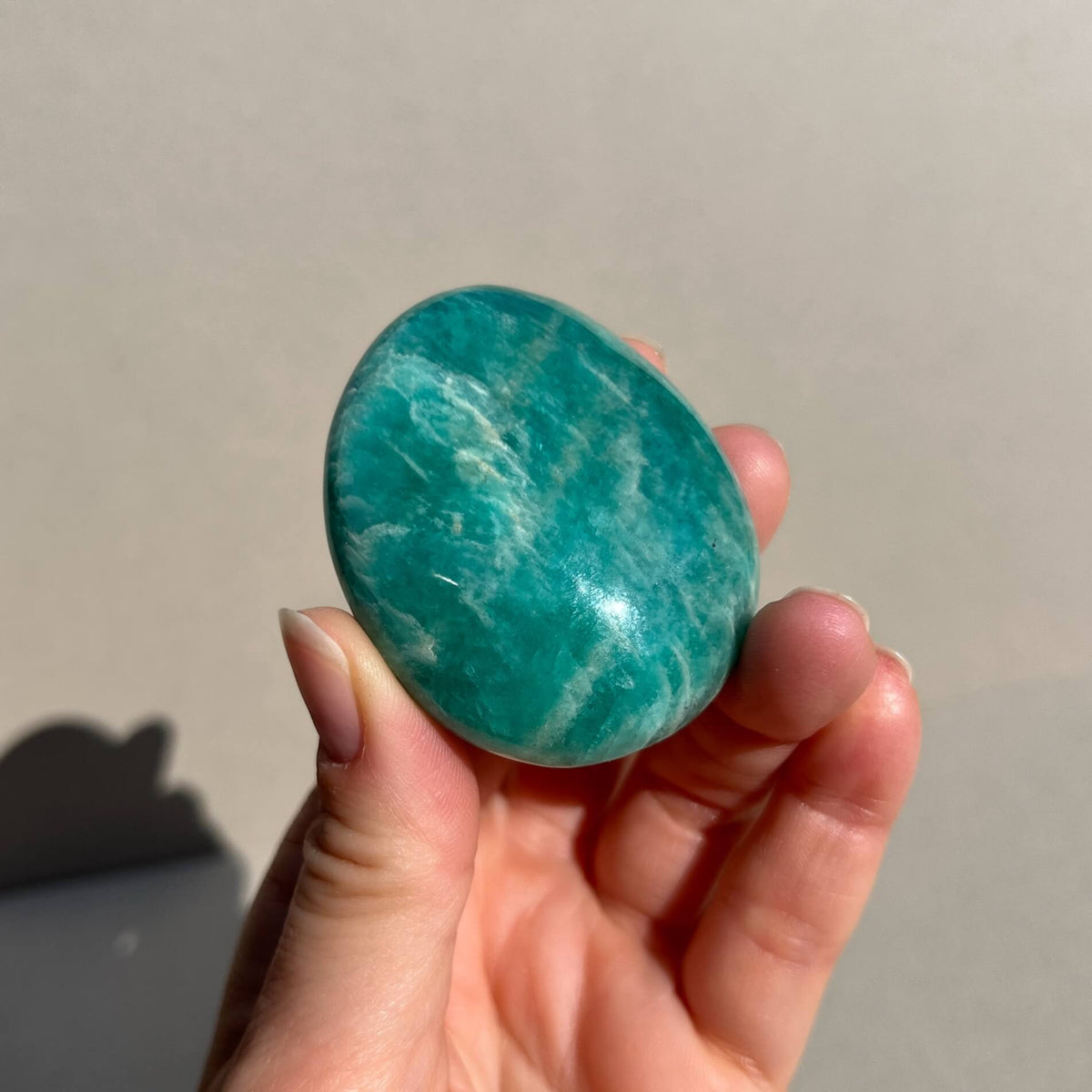A hand holding a polished Amazonite crystal palmstone with its characteristic teal and white banded pattern, set against a plain light taupe background.