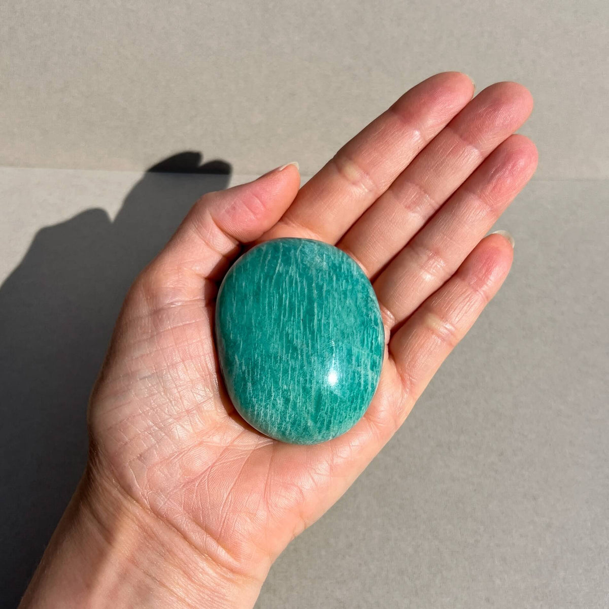 A polished oval amazonite palmstone in a vibrant turquoise-green colour with fine white striations, held in a person's open palm against a neutral grey background.