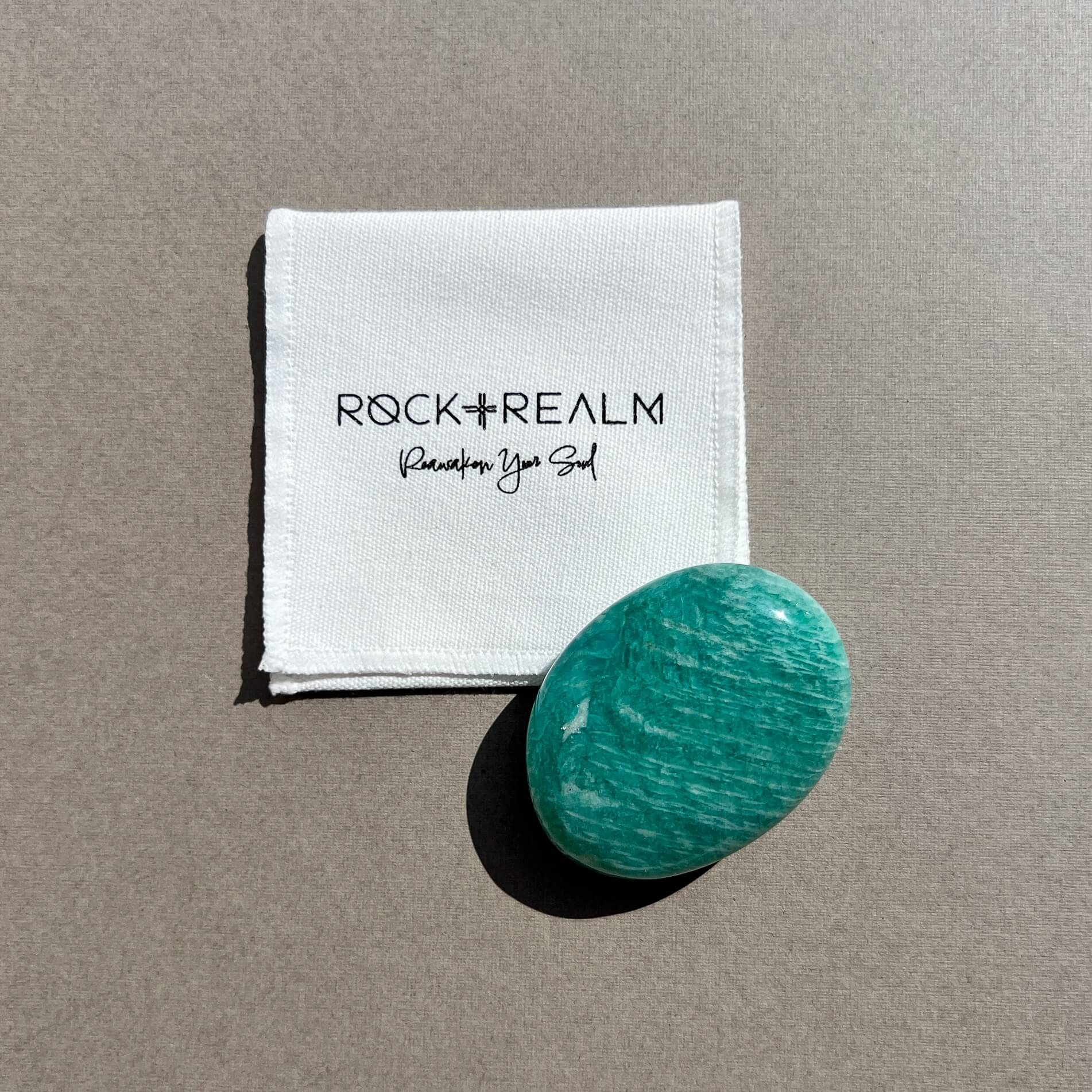 A polished oval-shaped turquoise-green amazonite crystal palm stone next to a white cotton pouch printed with the brand name 'ROCK + REALM' set against a neutral taupe background.