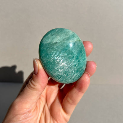 A hand holding a polished green Amazonite crystal stone with an iridescent sheen against a plain grey background.