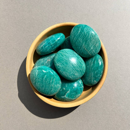 A bowl of polished green amazonite palmstones on a neutral grey background.