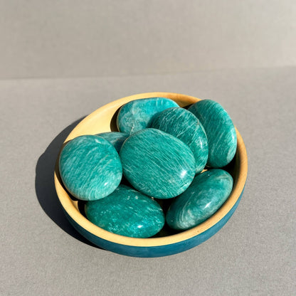 A collection of polished turquoise amazonite palm stones arranged in a small wooden dish against a neutral taupe background