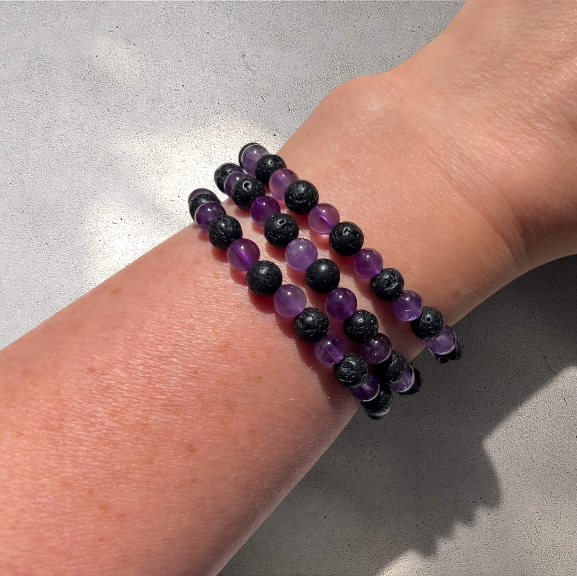 Stack of 3 black lava and purple amethyst crystal bead bracelets on a wrist against a neutral background