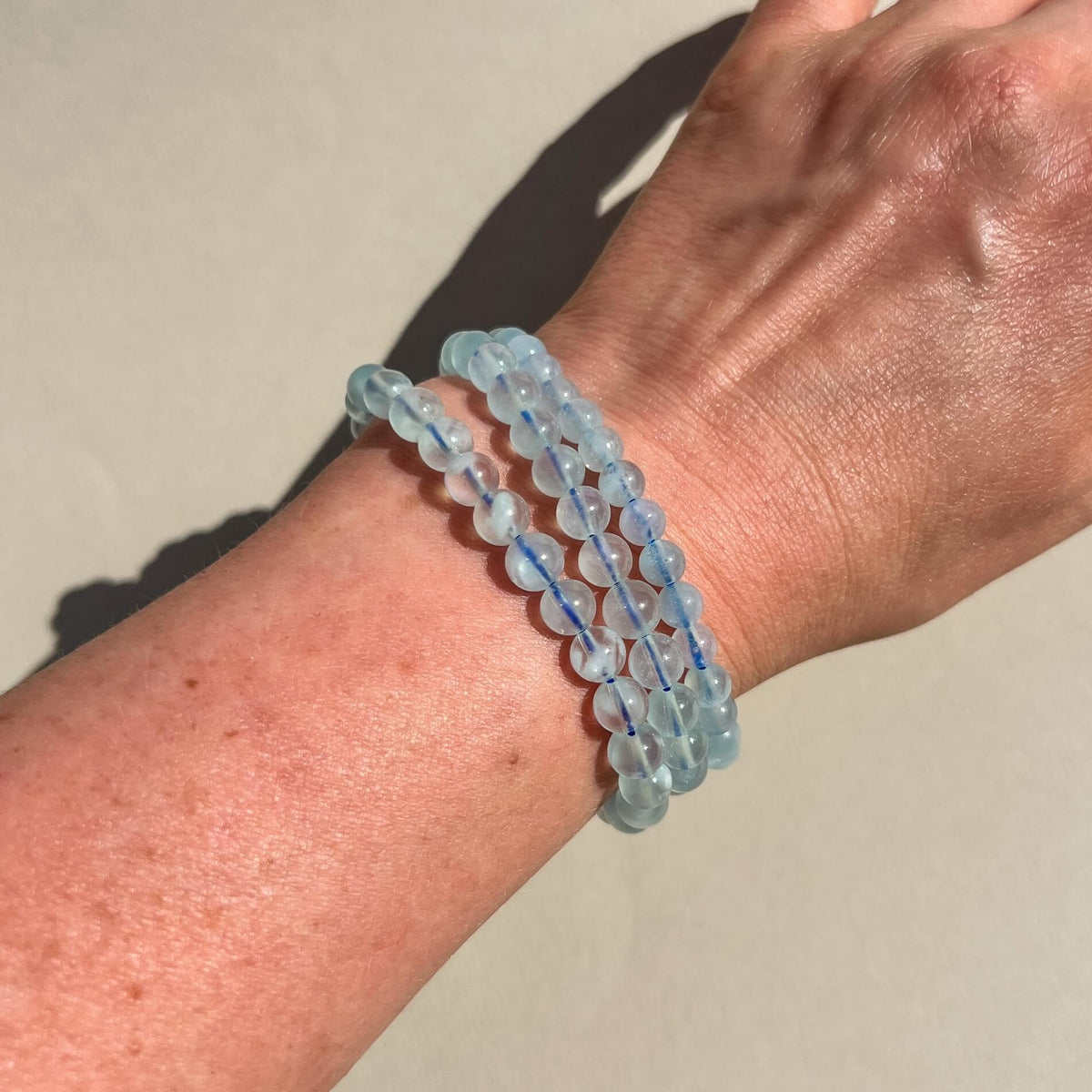 Three Aquamarine beaded stretch bracelets made of light blue marbled beads worn on a person's wrist, shown against a neutral beige background.