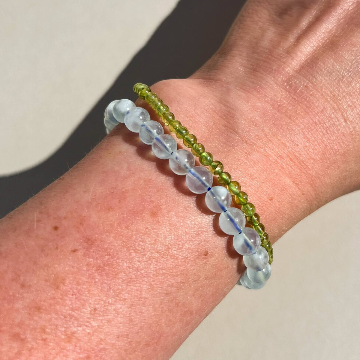 Two beaded bracelets worn on a wrist: one strand with translucent light blue Aquamarine round beads and another with small olive green Peridot round beads, shown against skin on a neutral light background.