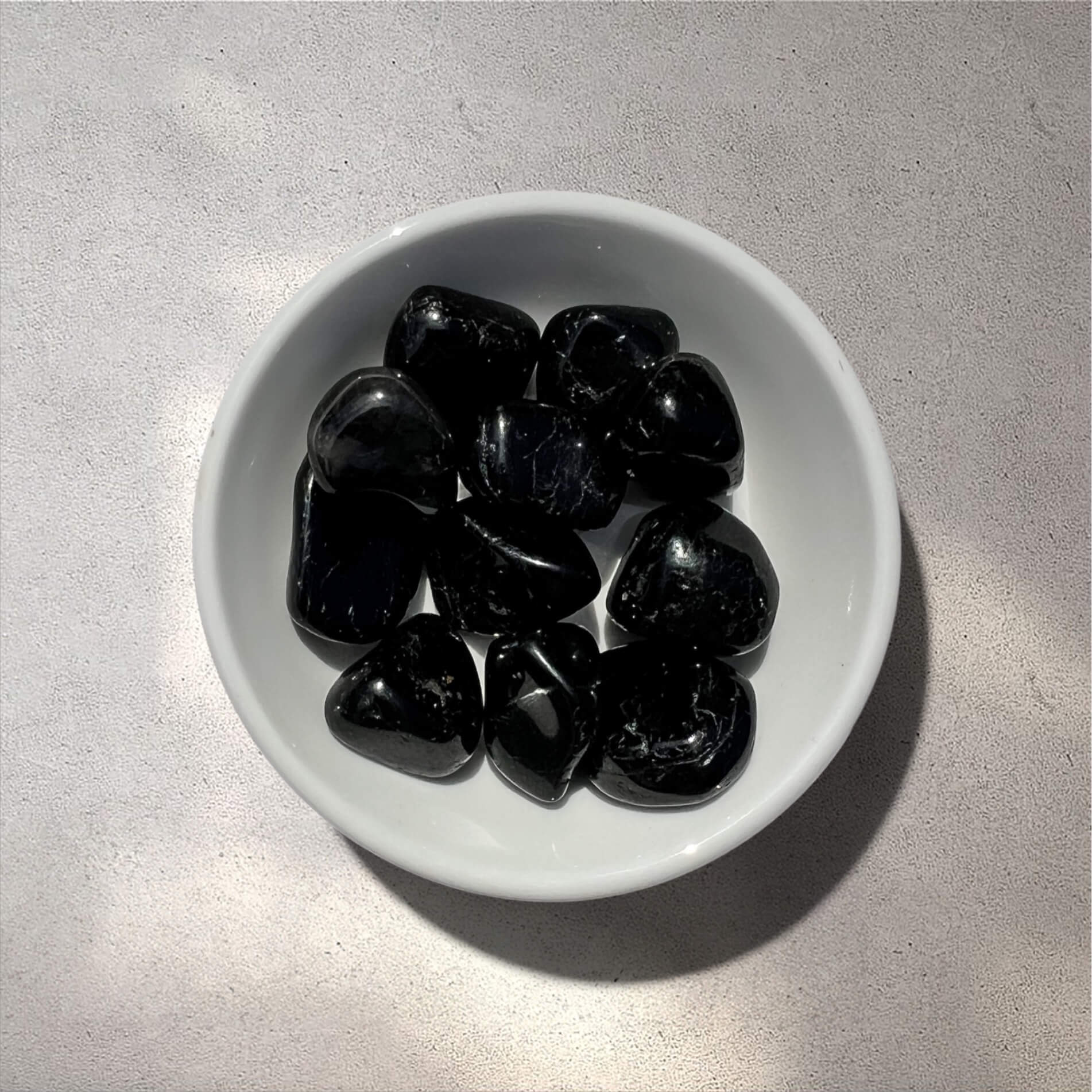 A bowl of polished black tourmaline tumbled stones on a light grey textured surface.