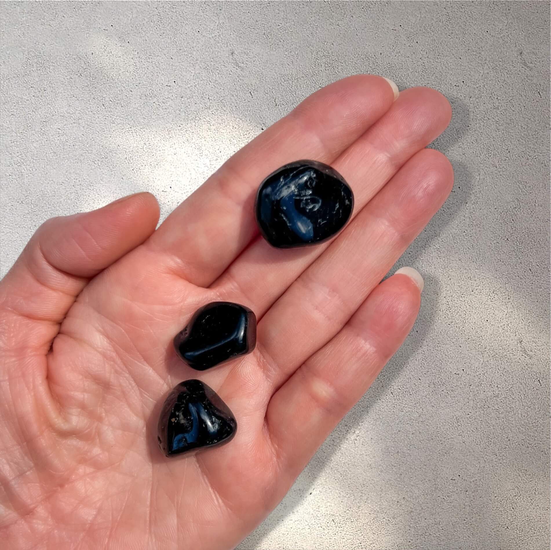 Three polished black tourmaline tumbled stones resting on an open hand against a light gray textured background.