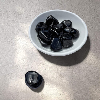 A white ceramic bowl filled with glossy black tourmaline polished tumbled stones, with one matching stone placed beside it on a light textured surface.