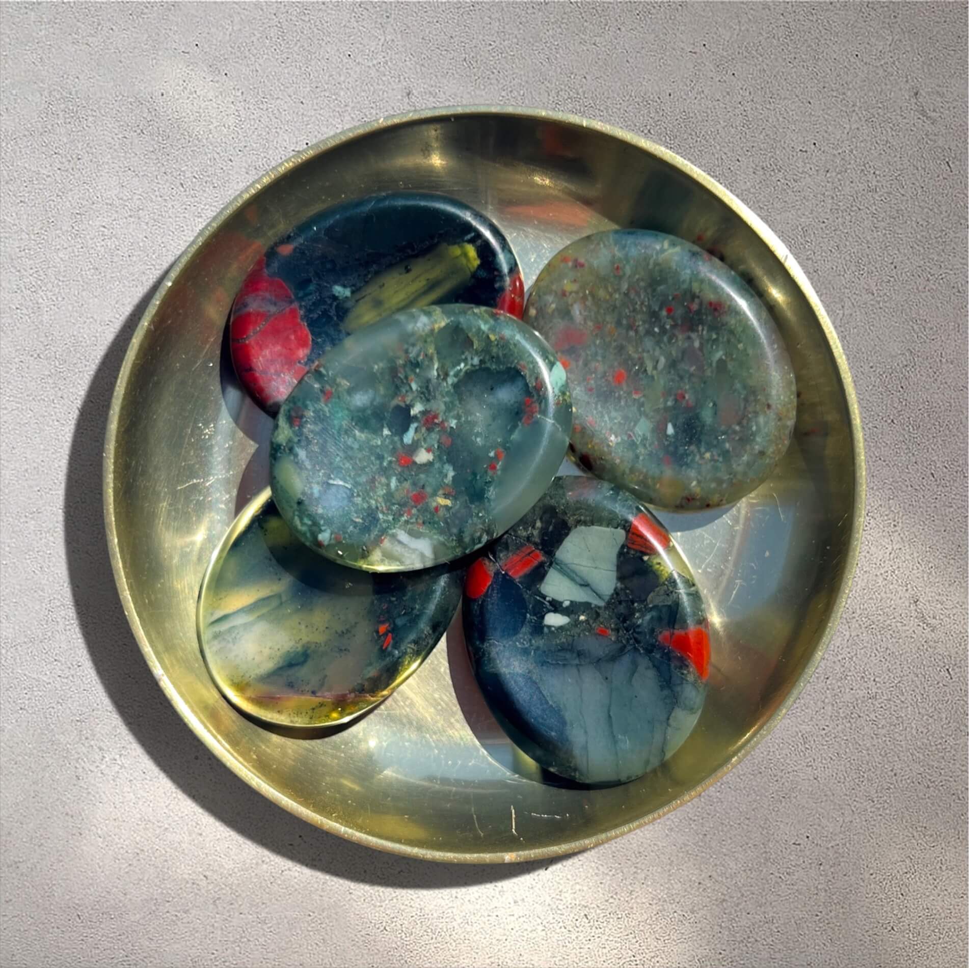 Five polished bloodstone worry stones with marbled green and red patterns arranged in a round brass tray on a light grey surface.