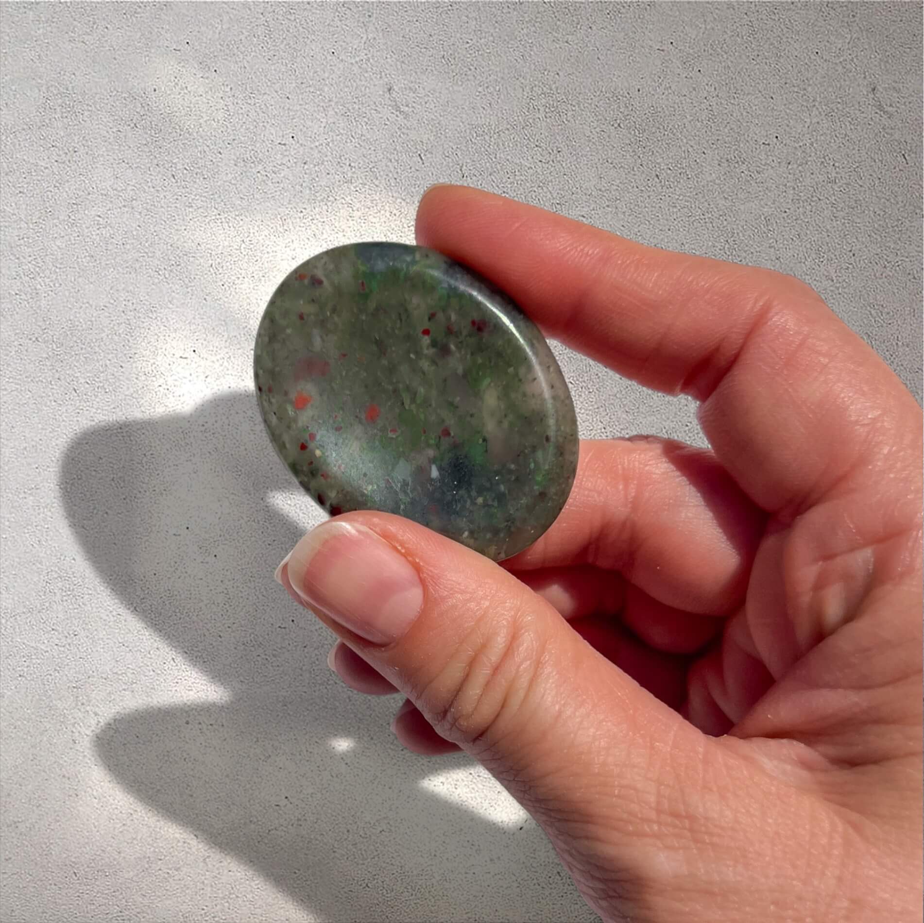 A hand holding a polished oval bloodstone worry stone with a mottled green appearance and small red flecks, set against a light grey textured background.