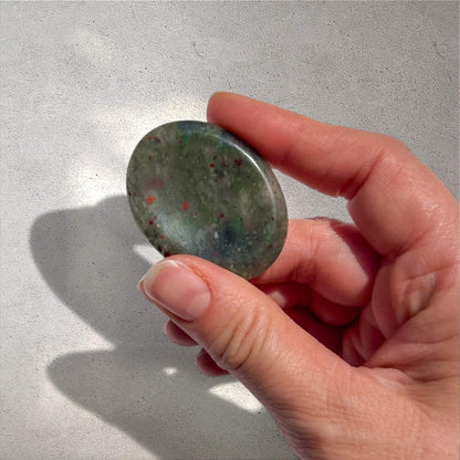 A hand holding a polished oval bloodstone worry stone with a mottled green appearance and small red flecks, set against a light grey textured background.