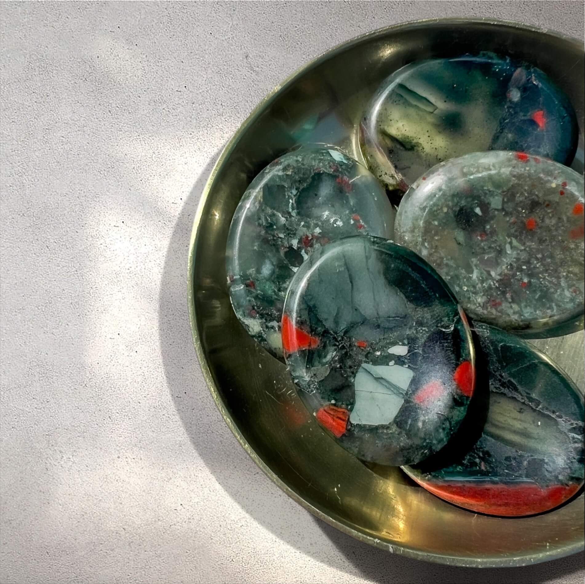 Five polished oval bloodstone crystal worry stones with green and red marbled patterns arranged in a round brass dish on a light grey textured surface.