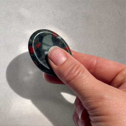 A hand holding a polished oval bloodstone  worry stone with a dark green base and red marbled inclusions, photographed against a light grey textured background.